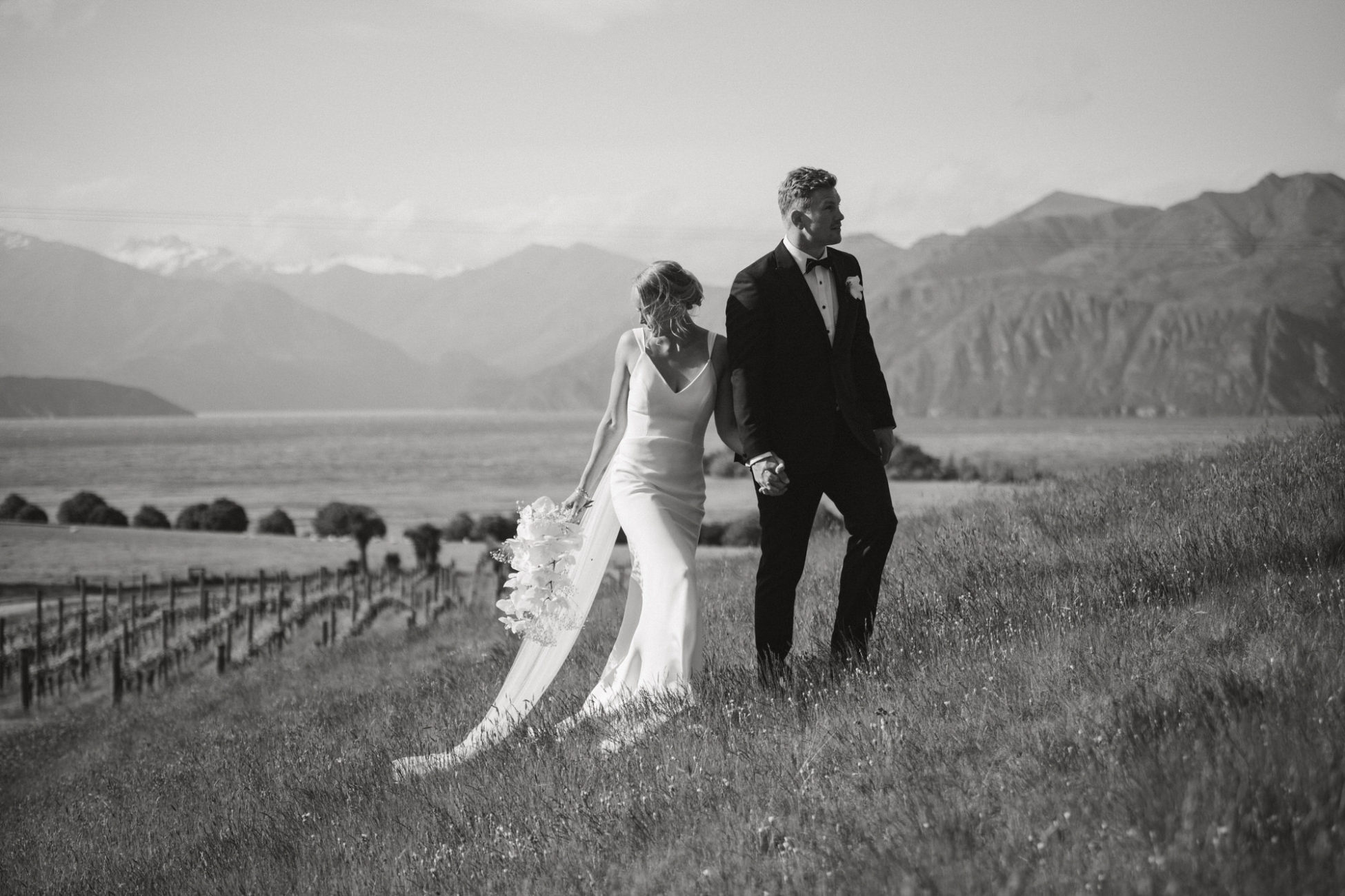 James and Alice holding hands and walking above the vineyard on their wedding day at Rippon Wedding venue in Wanaka